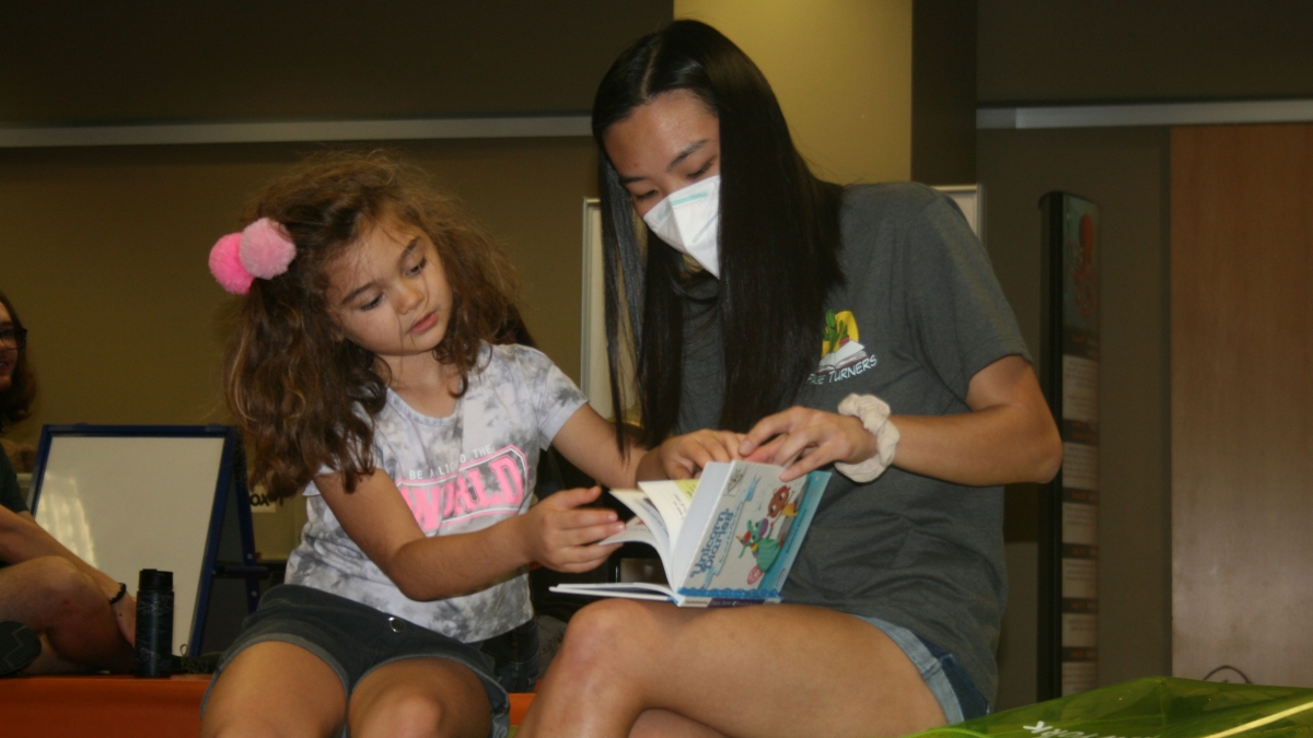 A college student wearing a mask read a book to a 7-year-old girl who is turning the pages