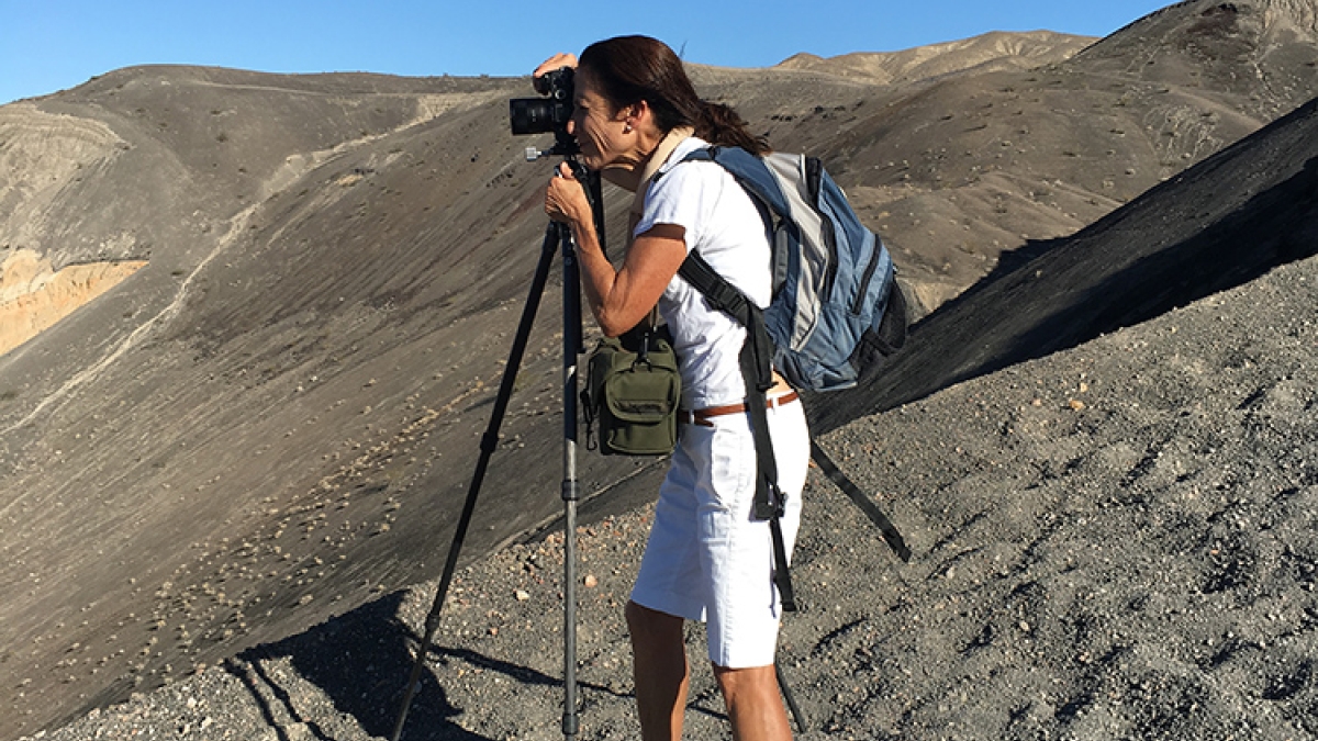 A dark-haired woman in white shorts and a white t shirt stands at a tripod on a gravel hill, taking a photograph