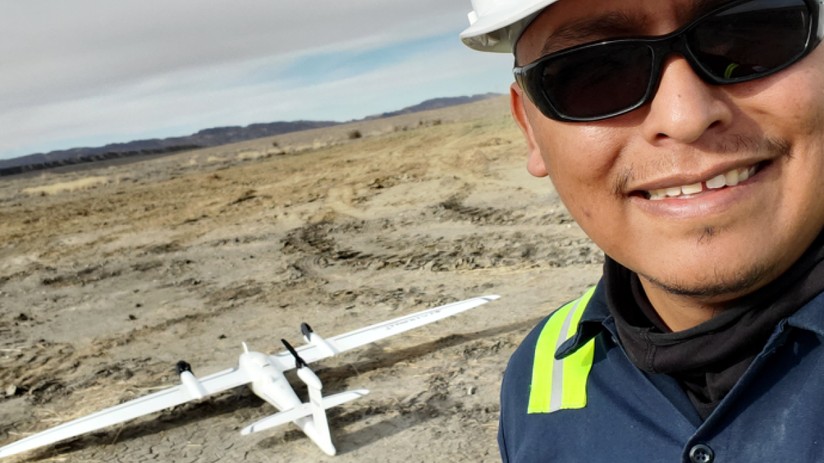 ASU student Preston Toehe wearing a hardhat and sunglasses while smiling and holding a remote control for a drone, pictured in the background, surrounded by a desert setting.