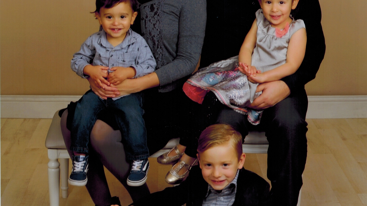 Rachel Hill, wearing a gray sweater and vivid pink lipstick, sits next to the right of her husband. They are each holding one of their children on their laps, and a third sits on the floor in front of them. They are posing for a family portrait.