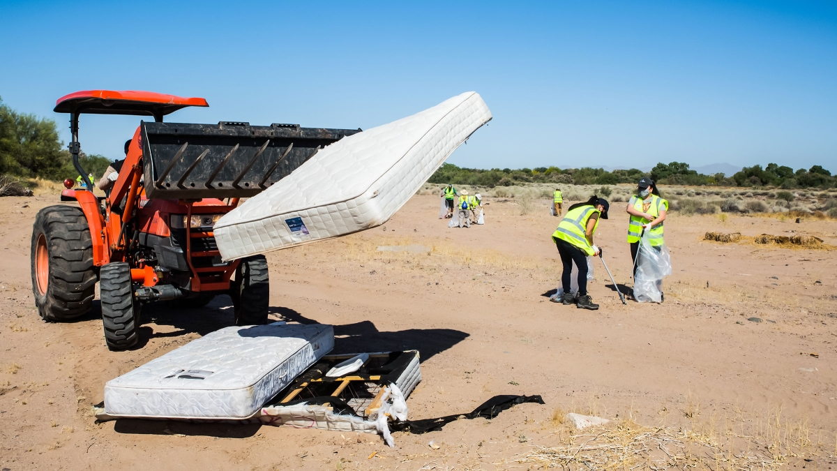 A forklift carries mattresses as volunteers in the background pick up trash on a dry riverbed
