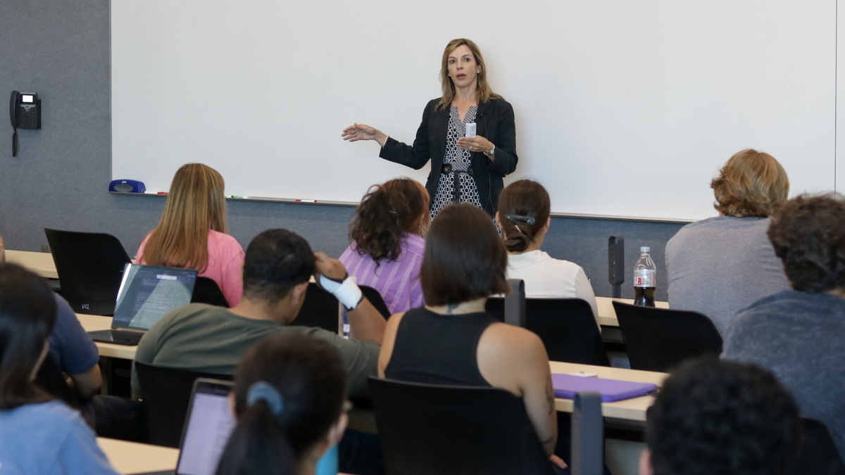 Woman standing at the front of a classroom speaking to people seated at tables.