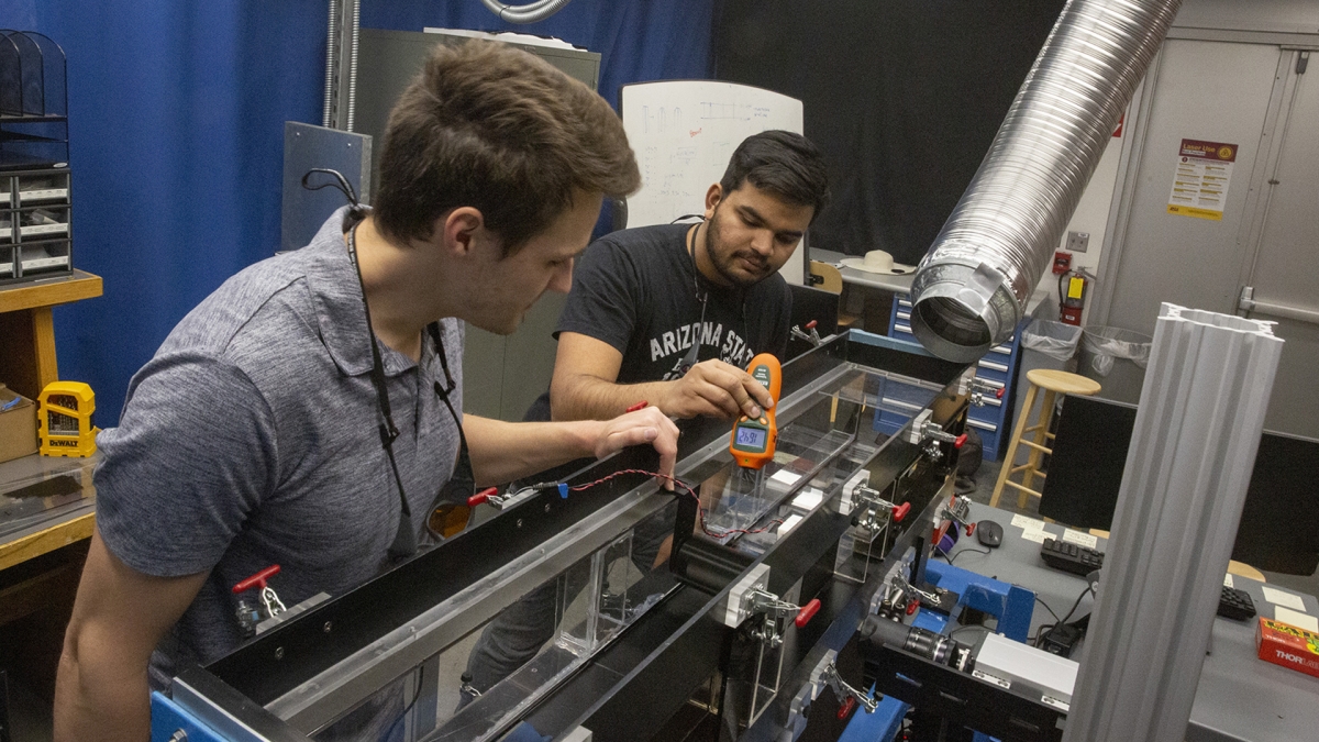 Two ASU students stand over a small wind tunnel as they insert a sample to test.