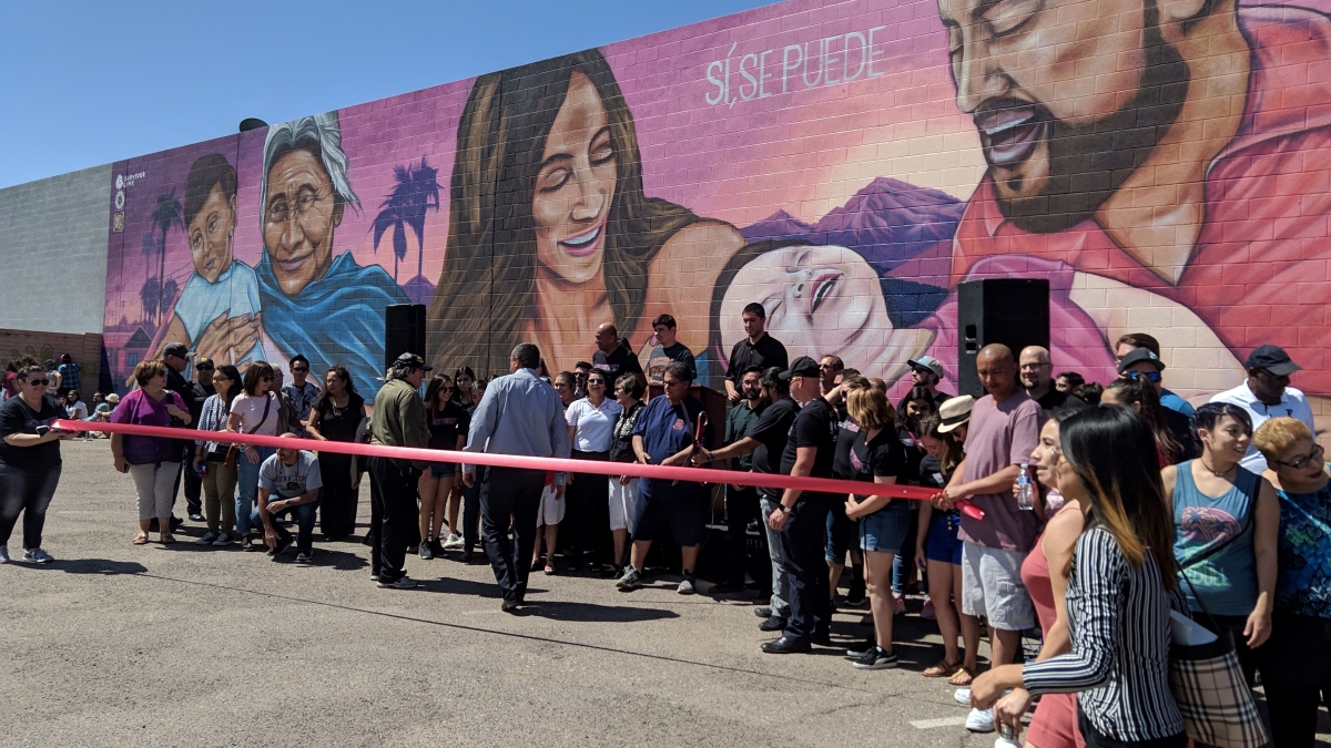 Community members gathered for a ribbon-cutting ceremony in front of a mural depicting a multi-generational Latino family.