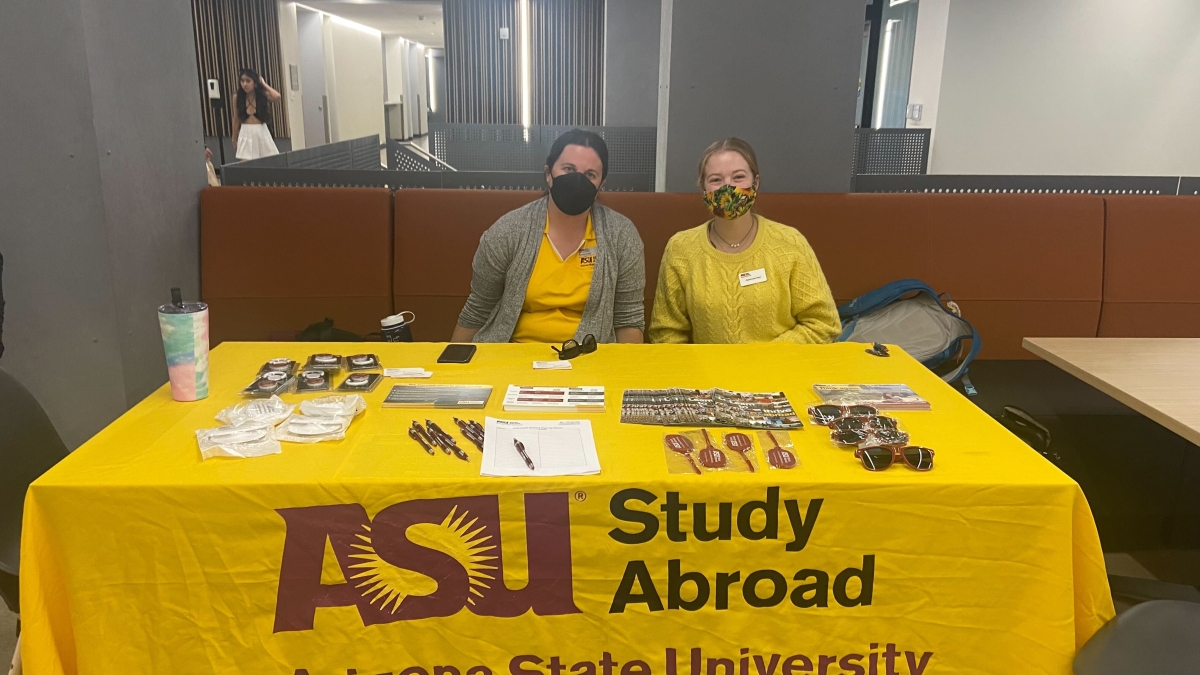 Two people sit behind a rectangular table covered in a gold tablecloth that has the logo for ASU Study Abroad on it. The table is covered in promotional items like sunglasses, pens and brochures. The two people are wearing gold and have on face masks.