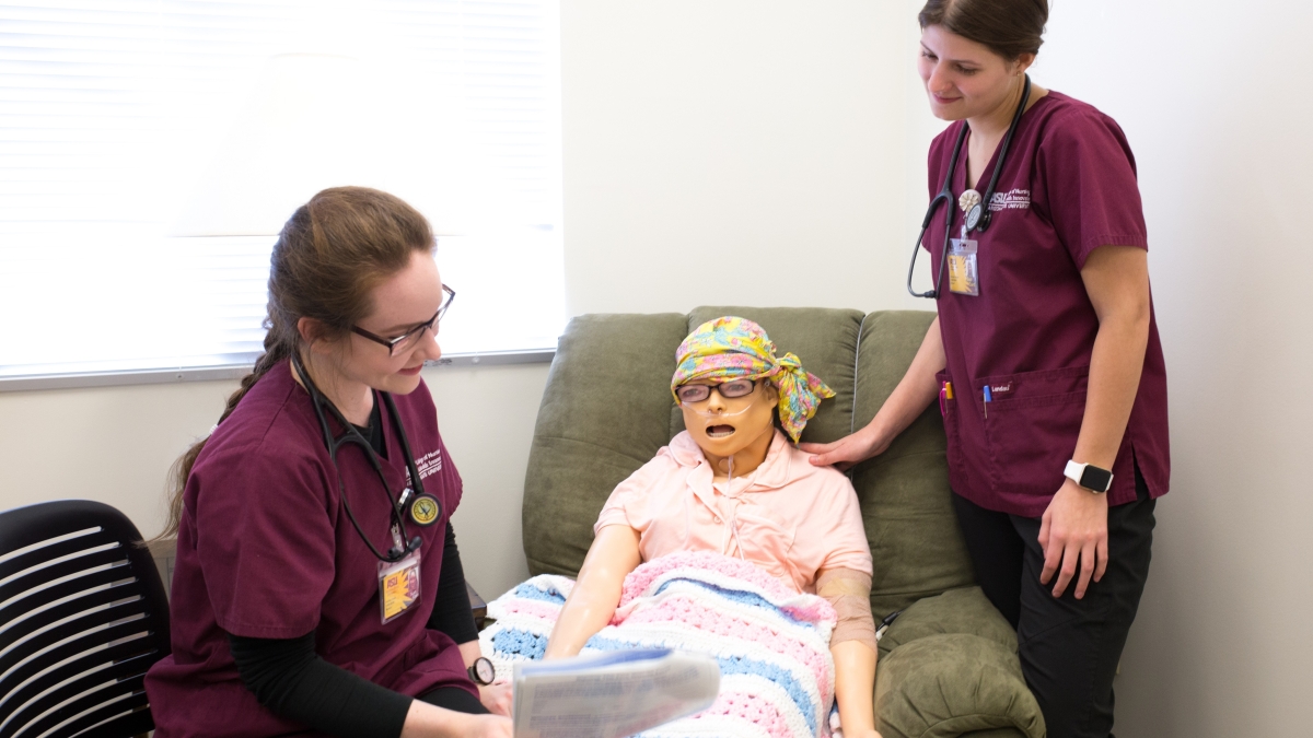 Two nursing students in maroon scrubs work through a simulation with manikin dressed as an older patient.