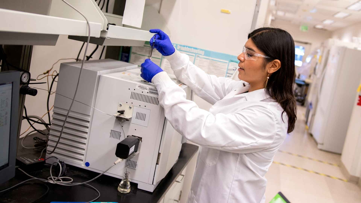 ASU student Aide Robles wearing a white lab coat, blue gloves and goggles in a research lab as she uses a large peice of machinery to examine a specimen.