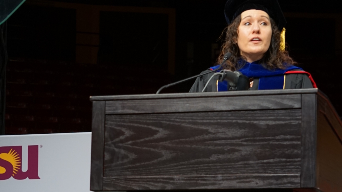 Woman standing behind a lectern wearing graduation regalia and speaking into a micorphone.