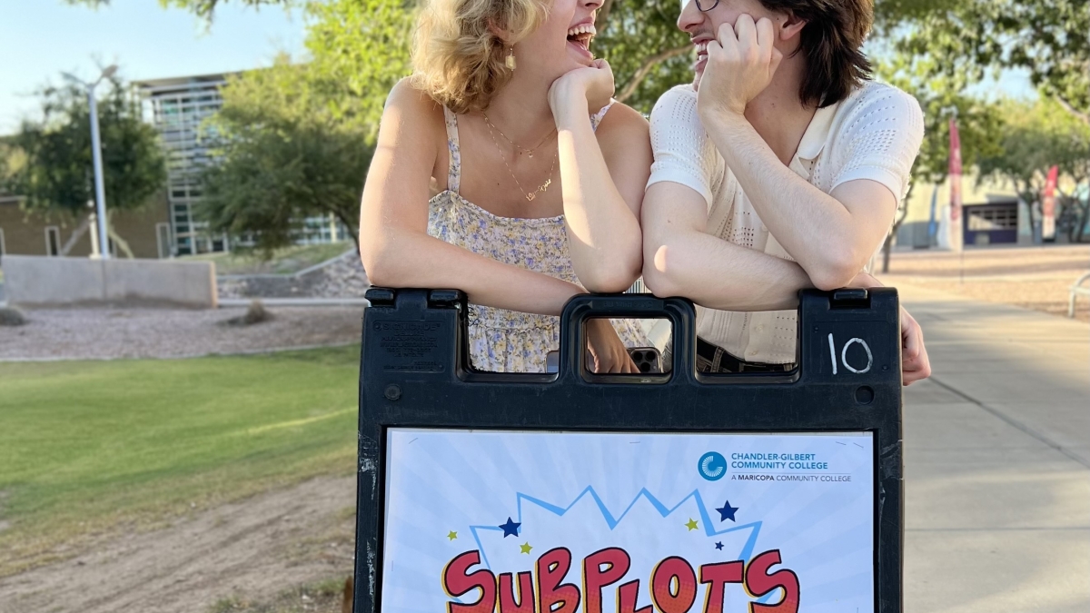 Two ASU alumni stand outside, leaning on an A-frame advertising their upcoming show "Subplots"