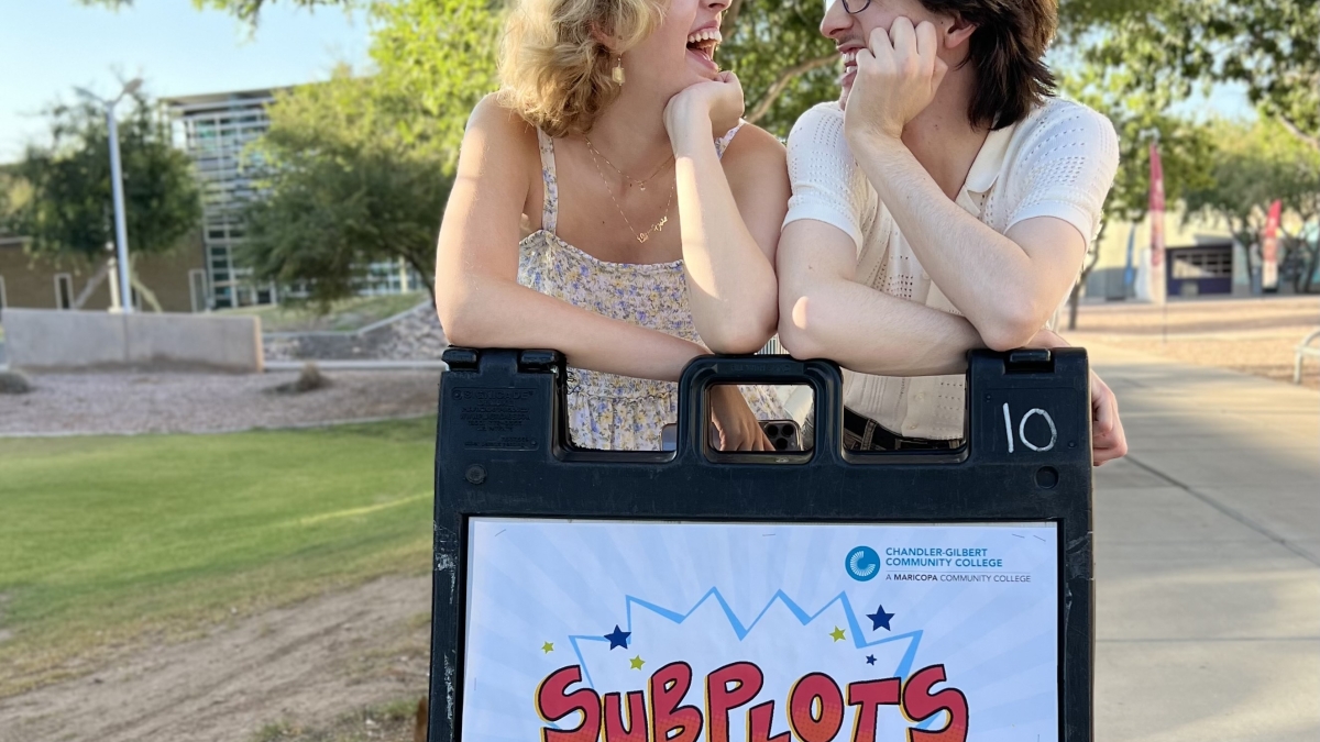 Two ASU alumni stand outside, leaning on an A-frame advertising their upcoming show "Subplots"