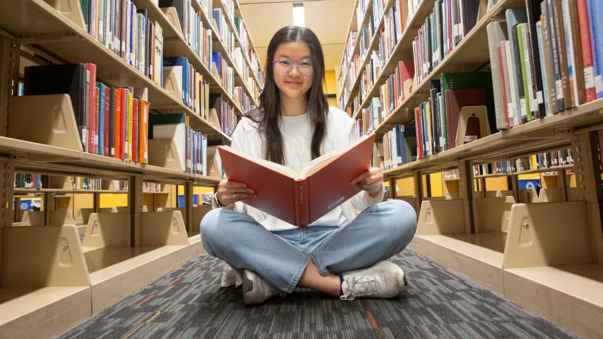 Student sitting on the floor between rows of books with book in their hand, looking at the camera.