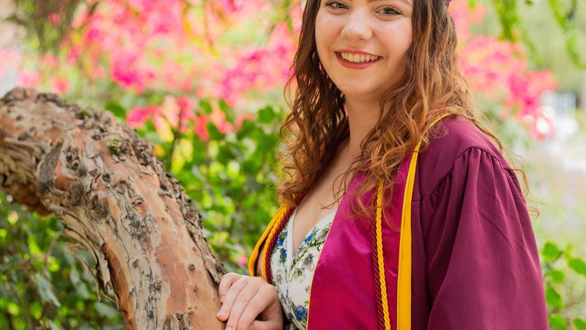 Sydney Campton poses in her maroon graduation robe over a light-colored top. Curly light brown hair hangs down from under her maroon graduation cap. Her hand is resting on a tree branch and there are out of focus leaves and flowers behind her.