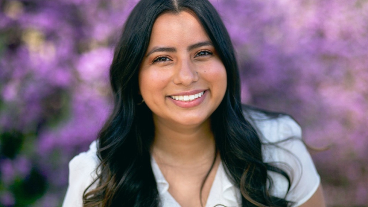 Portrait of Yazmin Reyes, a smiling young woman with dark hair and purple flowers in the background.