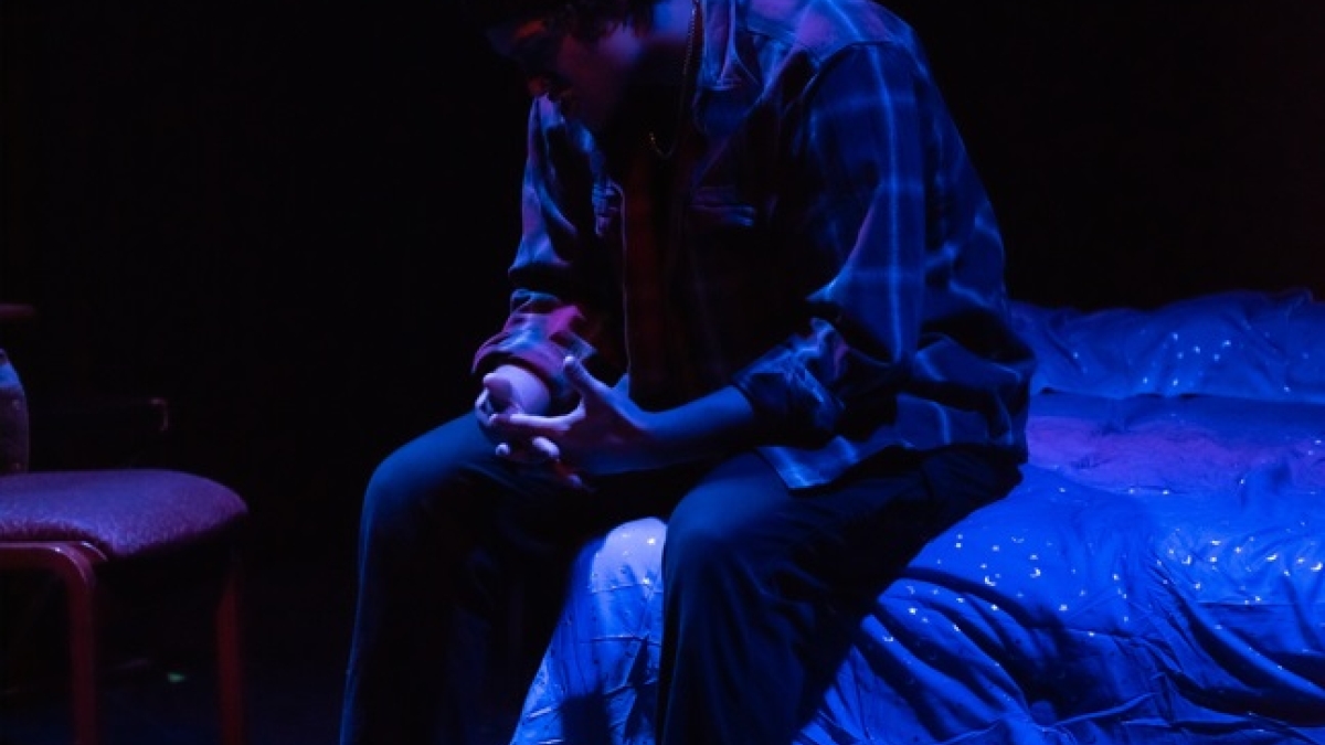 Man seated on a bed on a stage.