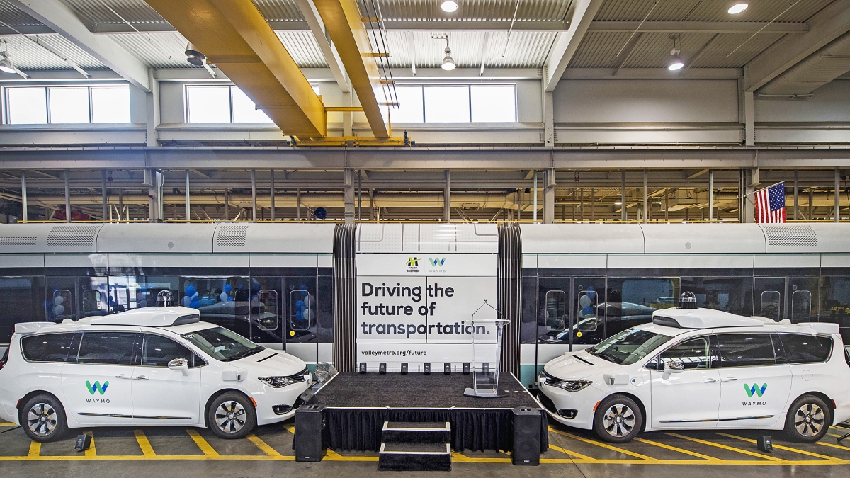 A small stage surrounded by Waymo autonomous cars and Valley Metro light rail train carriages