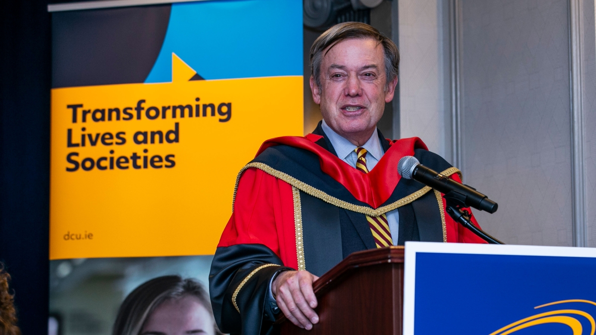 ASU President Michael Crow speaks behind a lectern with a DCU sign on the front.