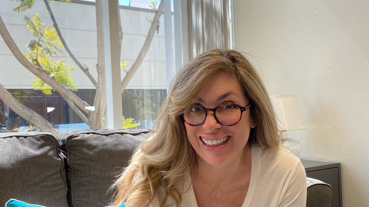 ASU alum Wendy Boring-Bray sits on a couch next to a window, in front of a pillow that reads: "Smile." Boring-Bray is wearing glasses, jeans and a white T-shirt and is smiling broadly.