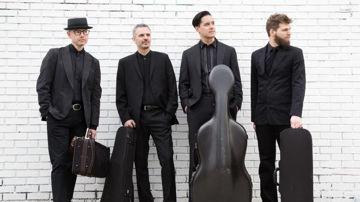 Four musicians in black suits stand against a white brick wall