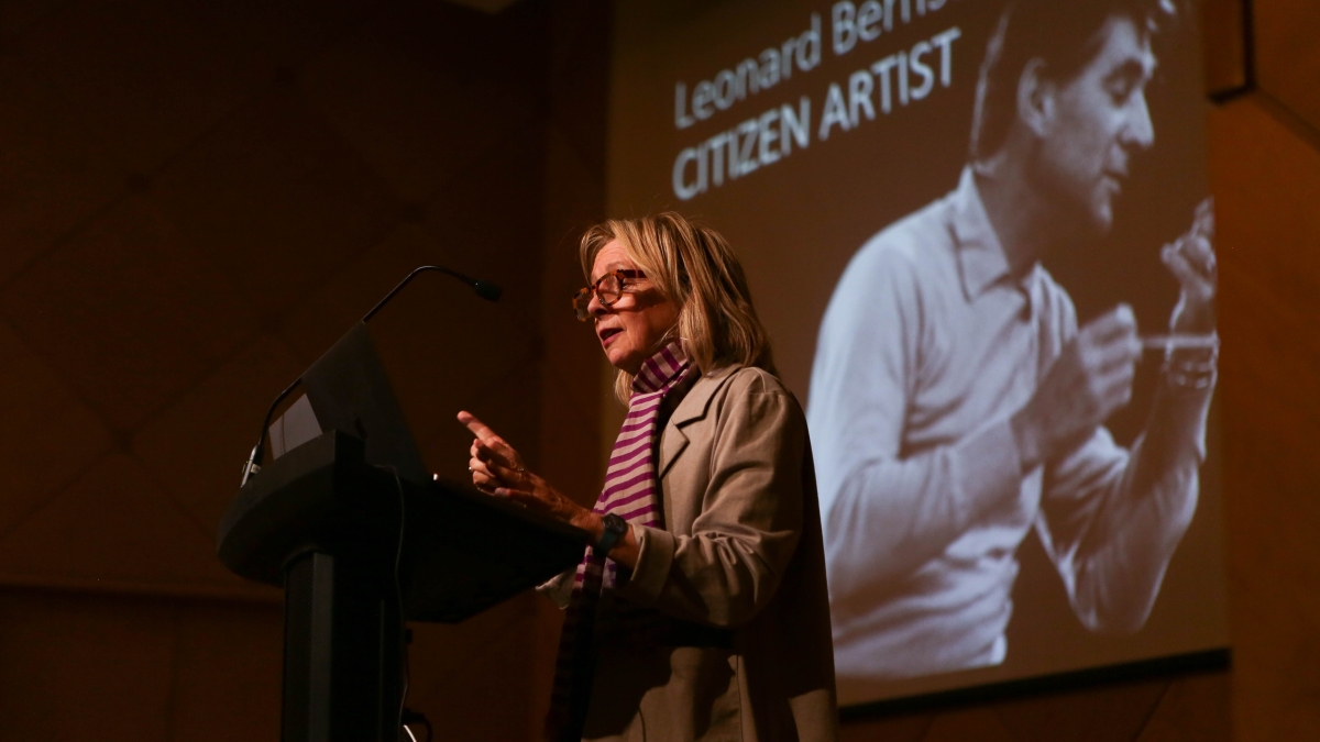 Woman on a stage speaking into a microphone in front of a screen showing a photo of Leonard Bernstein.
