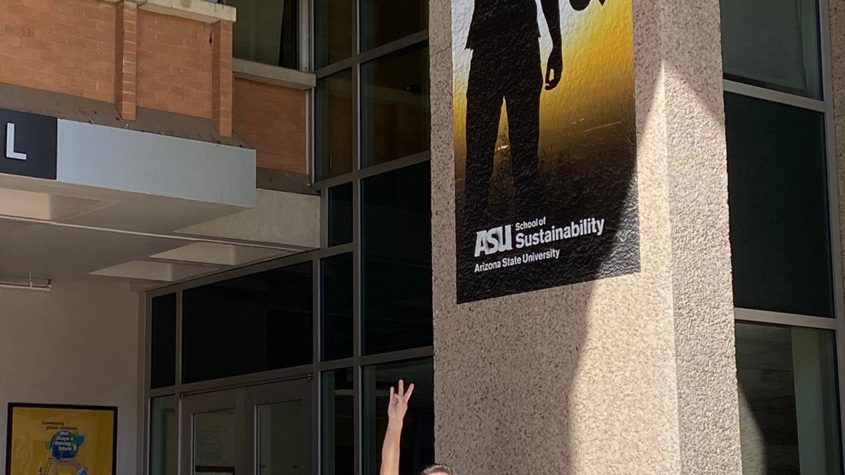 A man stands outside holding his hand in a pitchfork gesture, mimicking an ASU marketing sign above him.