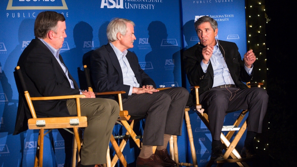 Three men discuss universal health care on a stage.