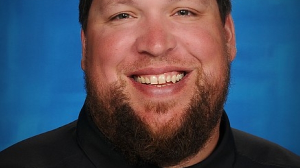 Portrait of ASU alum Colin Donovan. He has short black hair and a beard and smiles at the camera in front of a blue backdrop.hirt