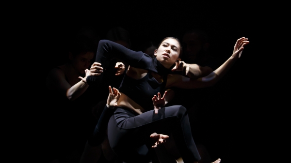 A dancer wearing all black is shown mid-performance on a dark stage.