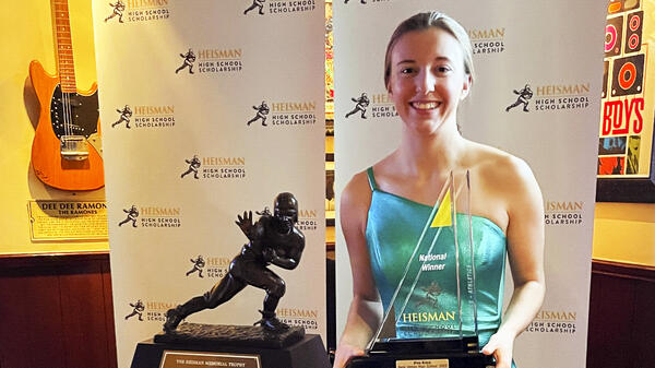 Viva Kreis hold award standing in front of Heisman High School Scholarship banners.