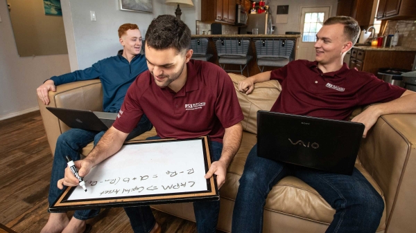 Three roommates sit on a couch working on laptops and a whiteboard