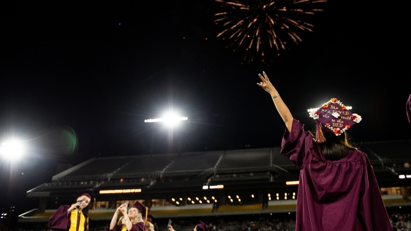 graduate in cap and gown and commencement making a pitchfork sign in front of fireworks