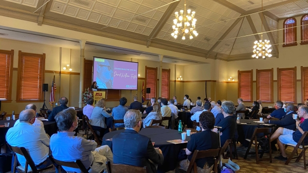 A small crowd of professors, alumni and other members of the ASU community sit at round tables facing a screen hanging on the wall. A person stands at a lectern in front of the screen.