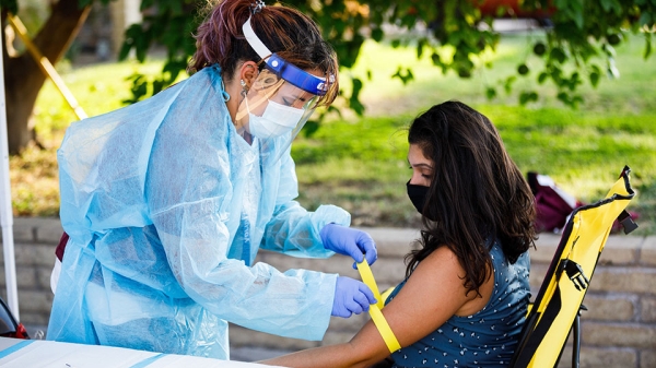 A woman in full personal protective gear prepares for a blood draw on another woman's arm.