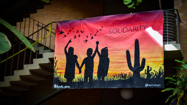 Photo of a mural hung from a staircase featuring silhouttes of three people holding hands in a desert landscape. The mural has the word "solidarity" printed on it.