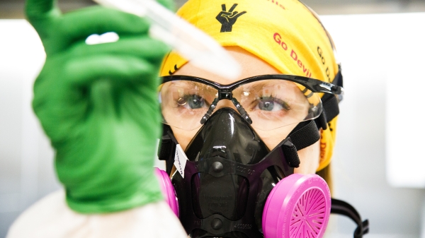 A researcher wearing a gold head cloth with ASU logos on it holds a test tube while wearing a gas mask