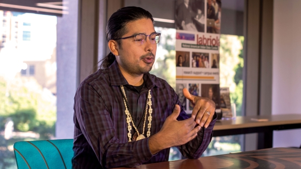 A Native American man wearing shell and bead necklaces and a plaid shirt gestures with his hands as he speaks while seated at a conference table, with a banner for the Labriola Center visible behind him