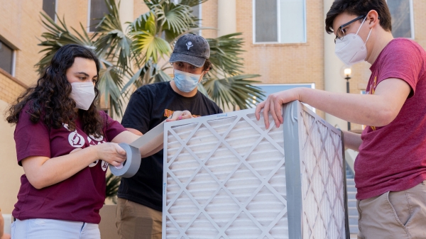 Three students wearing masks tape together air filters in the shape of a cube.