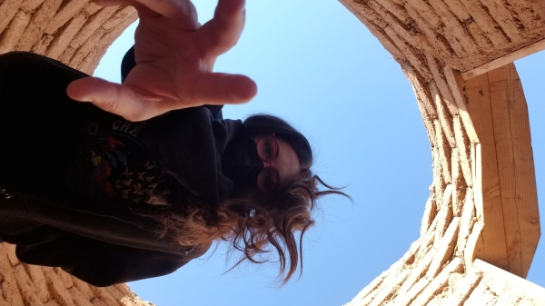 Self portrait of artist Celina Osuna. Osuna is picture from below, reaching toward the camera. Behind her is a blue sky and she is framed by bricks stacked in a cylindrical formation.