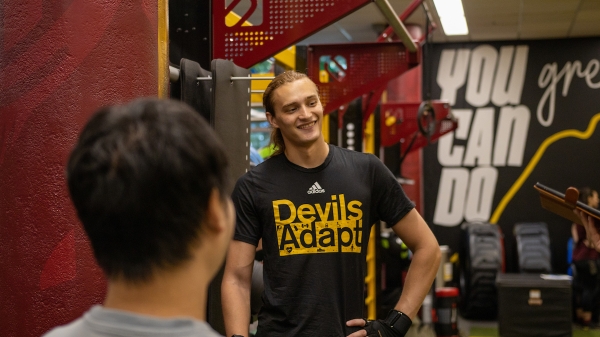 ASU student smiling and standing with hand on hip among weight training machines while wearing a shirt that reads "Devils Adapt."