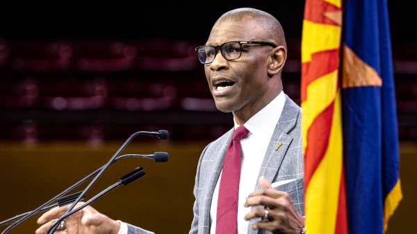 Man speaking into a microphone at a podium next to the Arizona state flag.