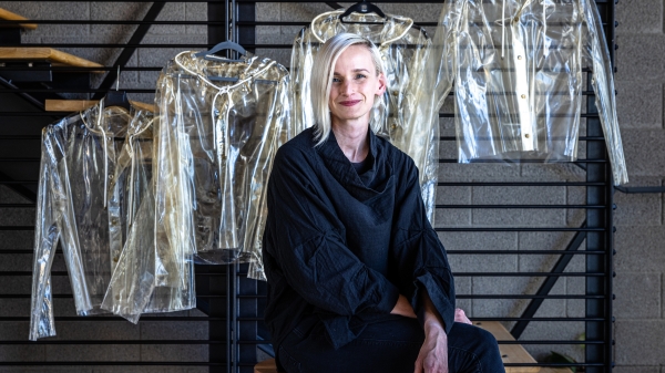 A woman with blonde hair wearing all black sits in a chair with see-through raincoats made of algae polymer hanging on a rack on the wall behind her.