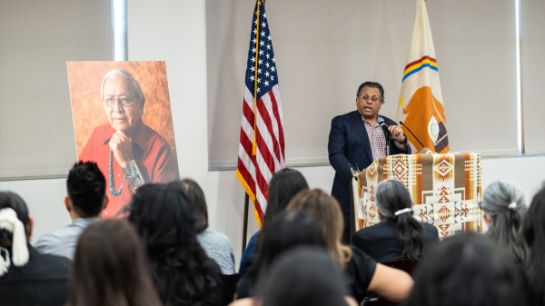 A man speaks at a lectern covered in a Native American textile with a memorial photo on an easel to his right