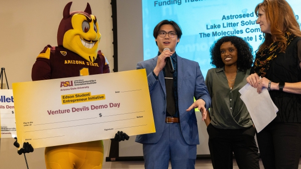 Three people stand next to ASU mascot Sparky holding a check as one speaks into a microphone.