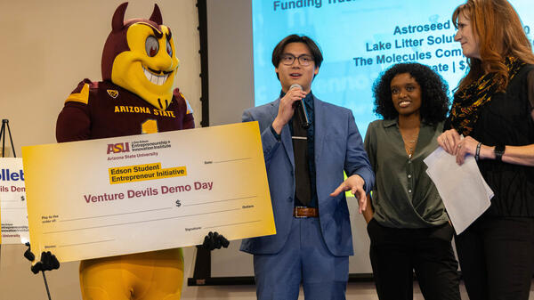 Three people stand next to ASU mascot Sparky holding a check as one speaks into a microphone.