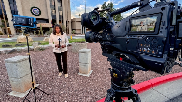 Linda Williams stands in front of a building with a microphone, with camera and film equipment around her.