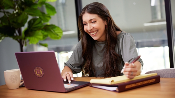 Woman working on laptop at table, smiling, sporting an ASU laptop sleeve, mug and binder. 