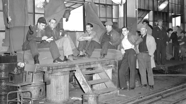Black-and-white photo of workers, some sitting and some standing, at an industrial plant.