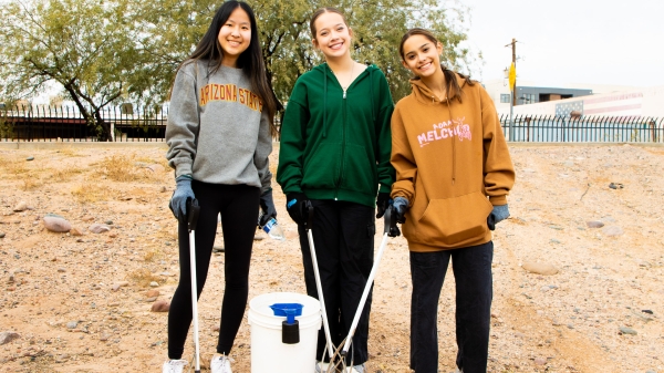 Students pose for a group photo holding trash pickers by a bucket in an outdoor setting.