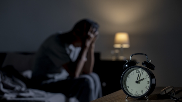 Man sitting on a bed with his head in his hands as a clock in the foreground shows the time as 2 a.m.
