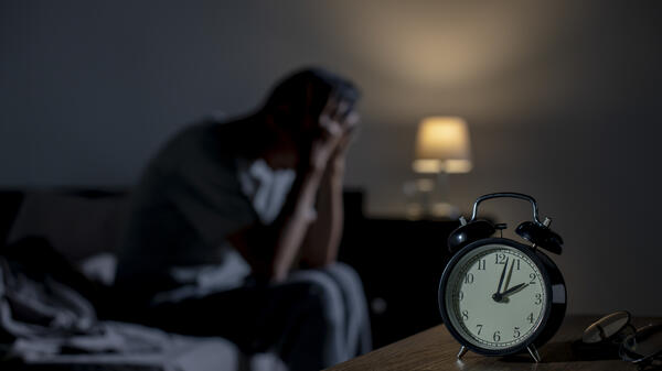 Man sitting on a bed with his head in his hands as a clock in the foreground shows the time as 2 a.m.