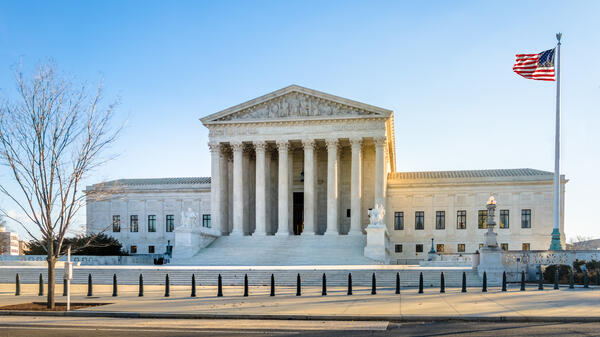 The U.S. Supreme Court building stands under a blue sky with the American flag waving.